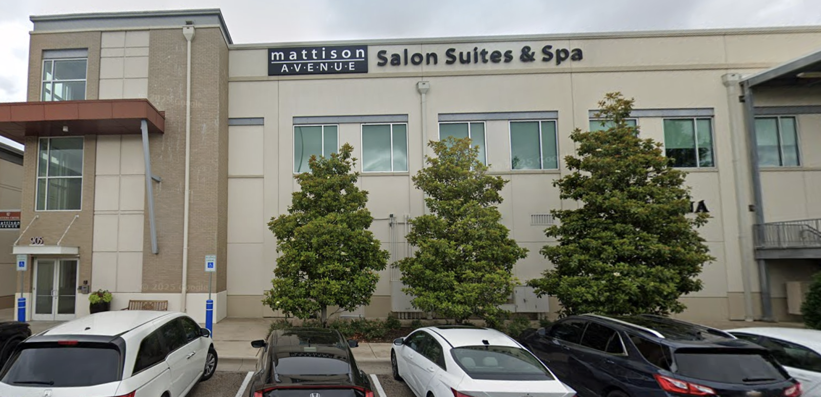 A modern building with "Mattison Avenue Salon Suites & Spa" signage, featuring a beige exterior, brick accents, and several trees in front. Multiple cars are parked in the foreground, and the sky is overcast.