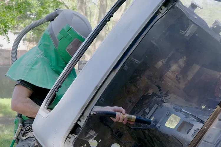 Technician uses abrasive blasting equipment to clean interior of vehicle panel, removing rust, paint, and contaminants to restore surface integrity and prepare for refinishing or protective coating application.