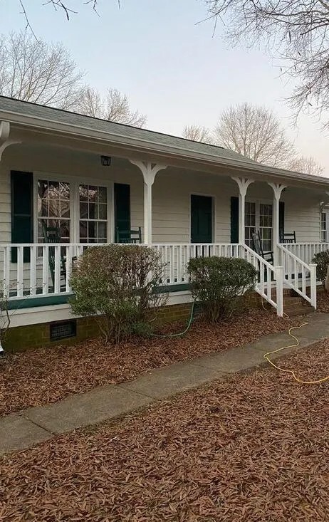 A single-story white house with a covered porch, green shutters, rocking chairs, and bushes in the front, surrounded by fallen leaves.