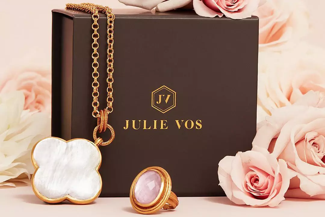 A sophisticated product visual by Tabula Rasa Social Stationers showcasing Julie Vos fine jewelry. The image features a gold-toned necklace with a distinctive white mother-of-pearl quatrefoil pendant and a matching gold-toned ring with an oval pink gemstone. Both pieces are elegantly displayed in front of a dark, branded Julie Vos jewelry box, complemented by a soft, rosy background, conveying luxury and classic elegance.