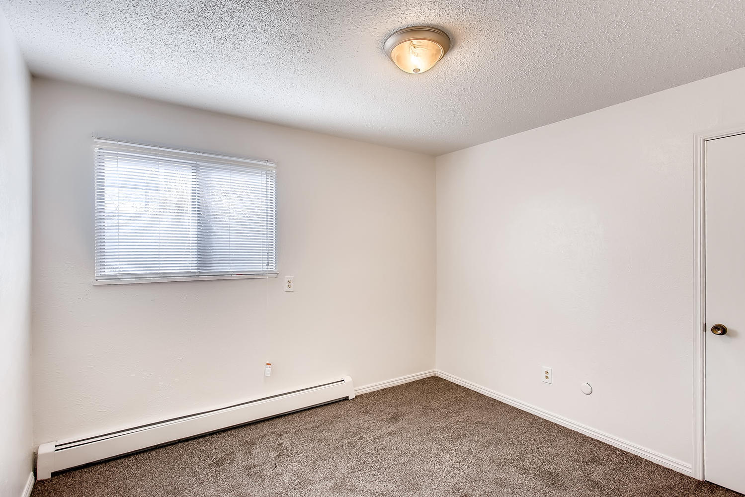 Carpeted bedroom with single light, single window, horizontal blinds, and door to bathroom.