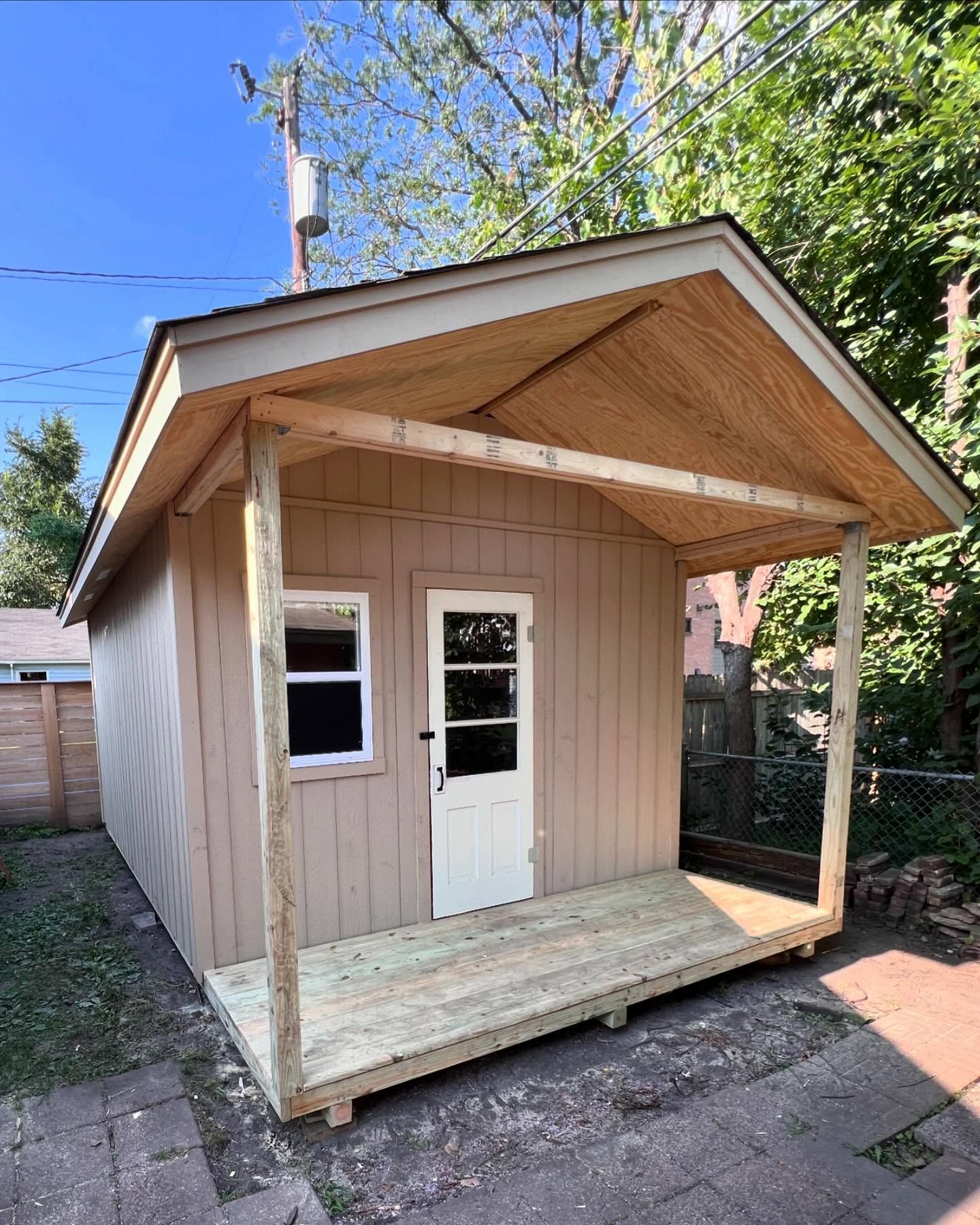 A newly built, light tan-sided storage shed features a covered front porch with a light-colored wooden floor and exposed plywood ceiling. The shed has a white door with a window and a separate small window on the wall.