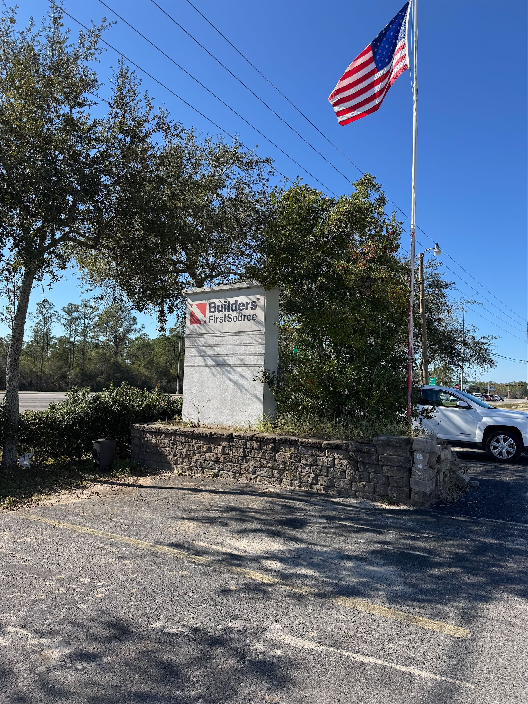 Builders FirstSource street sign in Santa Rosa Beach, Florida, featuring the company logo with an American flag flying proudly above, surrounded by trees and a parked vehicle.