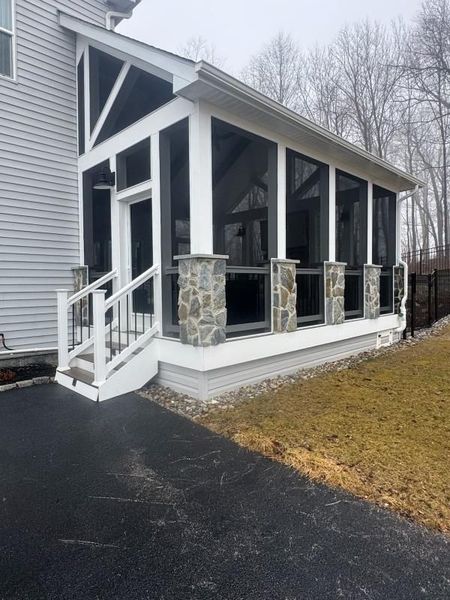 Enclosed screened porch attached to residential home with stone columns and stairs. The structure enhances outdoor living by providing sheltered space, combining construction and landscaping elements for comfort, durability, and aesthetic appeal.