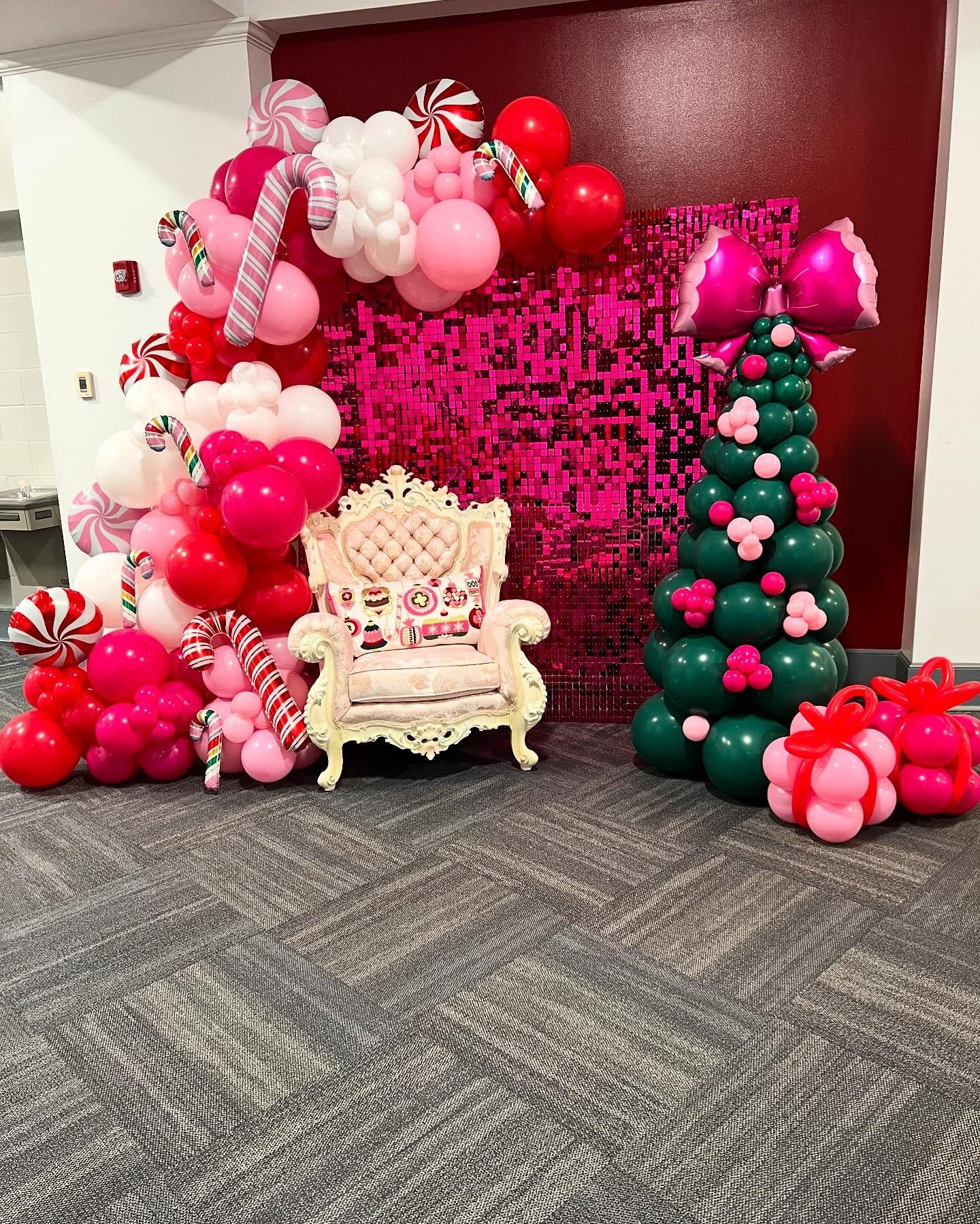 Festive holiday photo backdrop featuring pink and red balloon garland with candy cane accents, sequined pink wall, ornate chair, and balloon Christmas tree, creating a playful, cheerful indoor celebration setup.