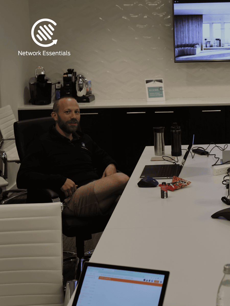A man sits at a table in an office, with two laptops and a coffee station in the background. The room has modern office furniture and a large screen mounted on the wall. The logo for "Network Essentials" is visible in the upper left corner.
