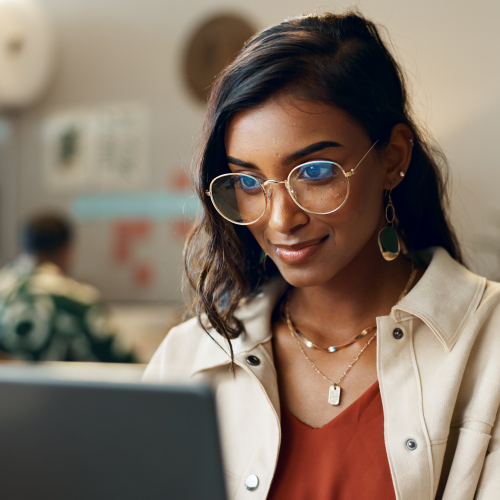 A young woman with long dark hair, wearing gold-rimmed glasses, layered necklaces, and earrings, smiles while working on a laptop in a cozy, modern workspace.