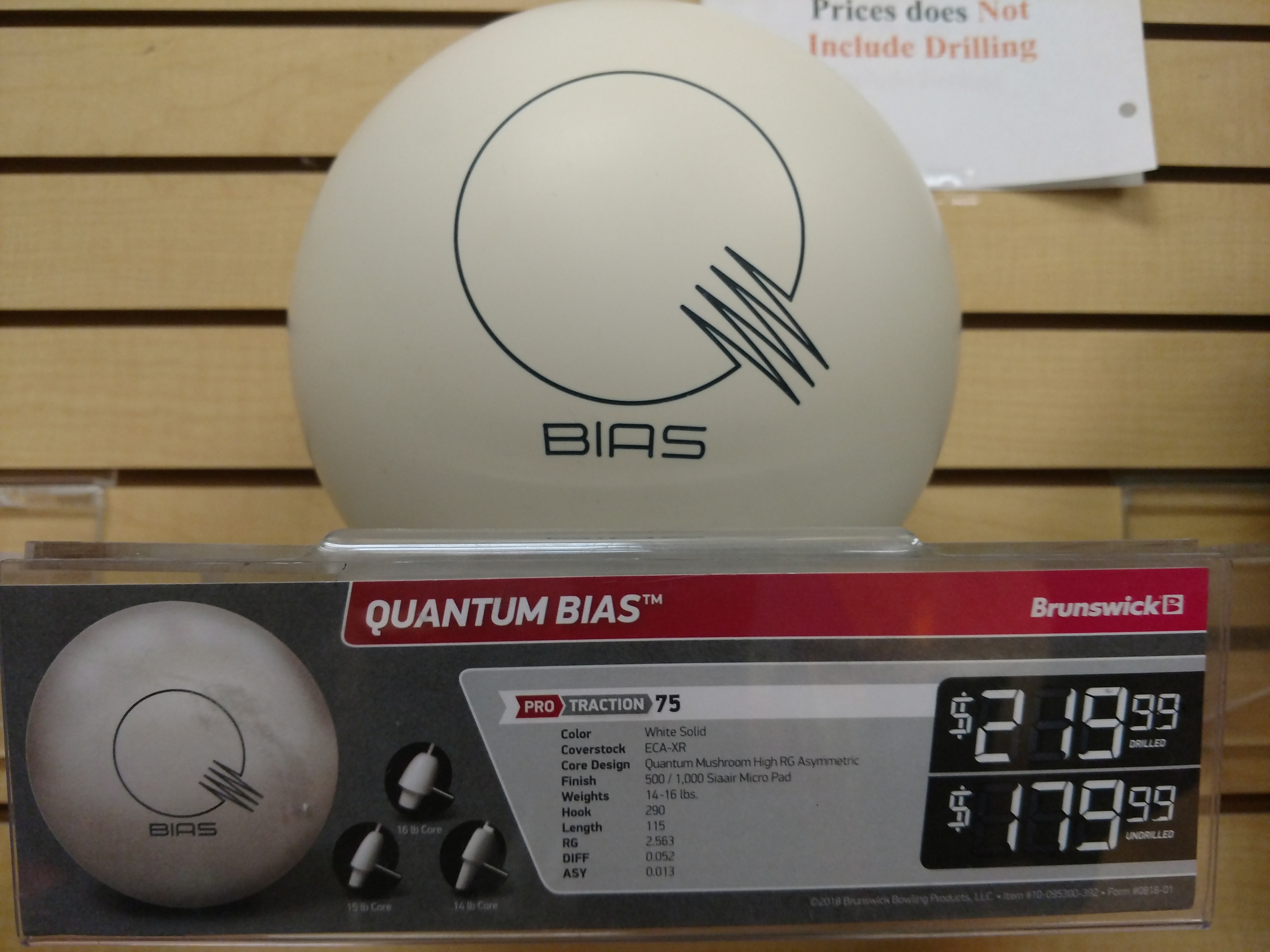 A white bowling ball with a black "BIAS" logo and a stylized Q graphic is displayed on a wooden shelf. Below the ball, a price tag shows "QUANTUM BIAS" with the Brunswick logo and prices for drilled and undrilled versions. Details about the bowling ball's specifications are also listed.