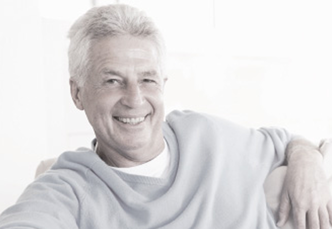 An older man with gray hair sits comfortably in a light-filled room, wearing a soft blue sweater and resting his arm casually.