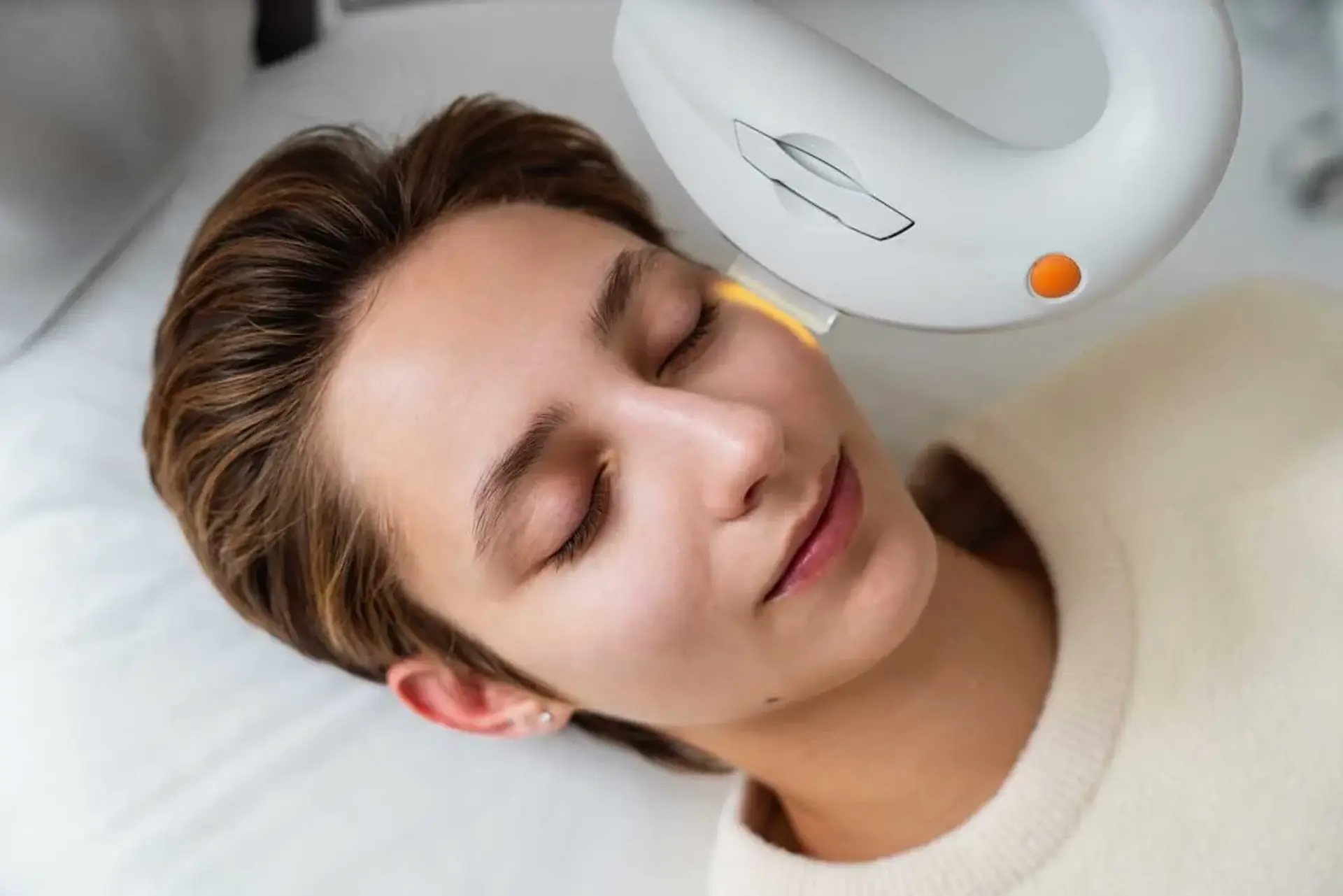 A person with closed eyes receives a cosmetic treatment on their face with a handheld device emitting a yellow light.