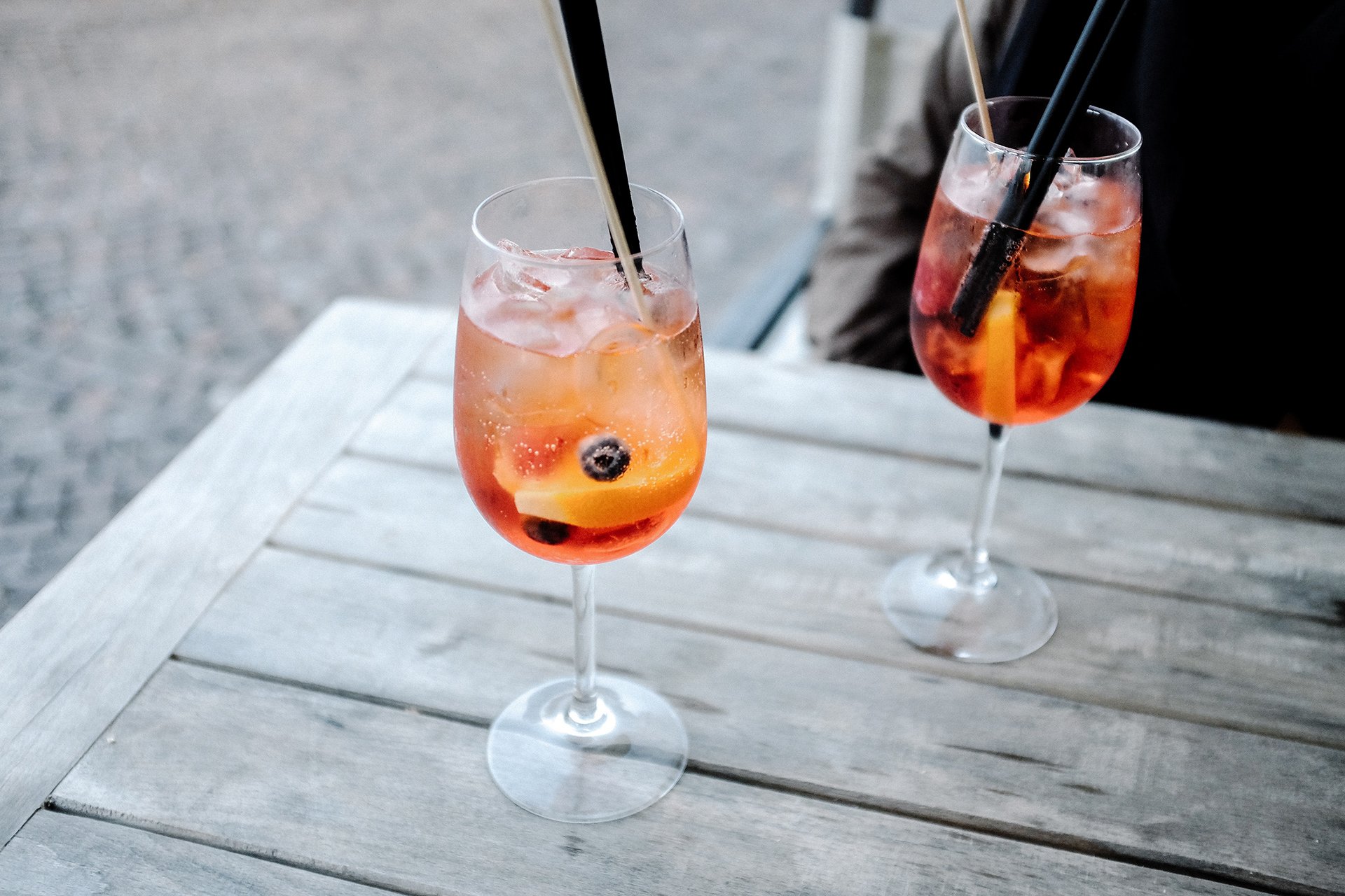 Two refreshing Aperol Spritz cocktails with ice, orange slices, and blueberries, garnished with black and wooden straws, are placed on a rustic wooden table outdoors.