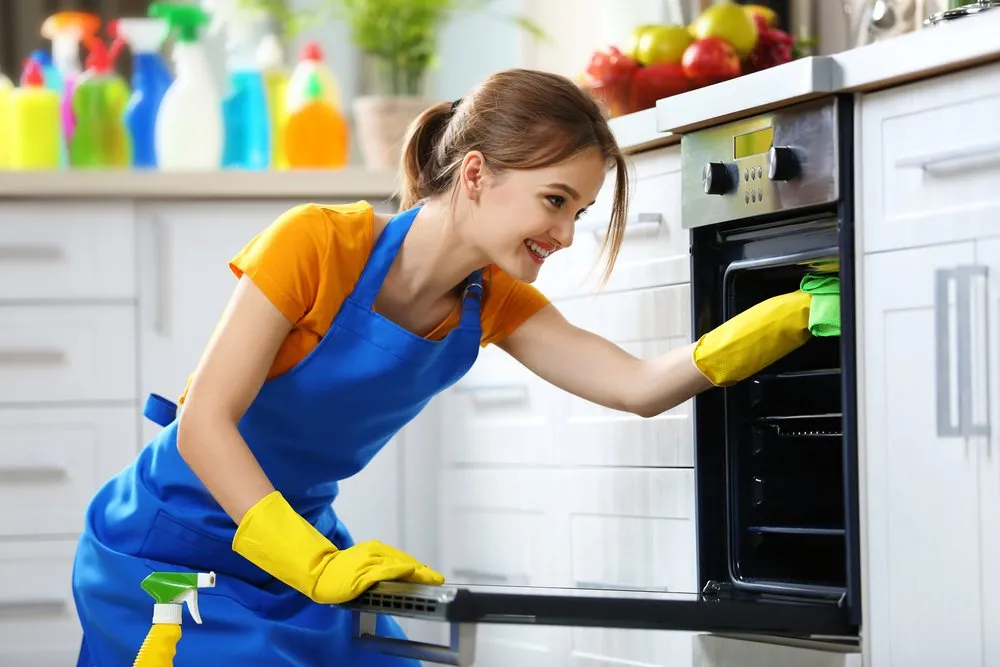 A woman wearing a blue apron and yellow cleaning gloves is cleaning the inside of an oven with a green cloth. She is smiling and appears to be focused on her task. In the background, there are cleaning supplies and fruit in bowls on a kitchen counter.