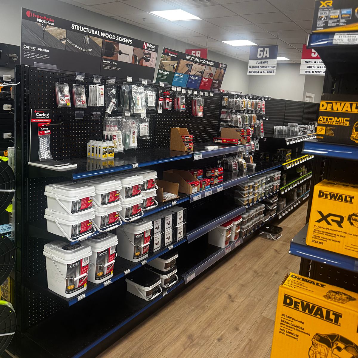 Wide view of a Builders FirstSource store aisle with black pegboard displays of structural wood screws, decking fasteners, adhesives, hardware, and white buckets of Cortex products. Overhead signs identify sections for structural wood screws, flashing, framing connectors, air hoses, fittings, nails, and screws. Yellow DEWALT boxes appear on shelving at the right.