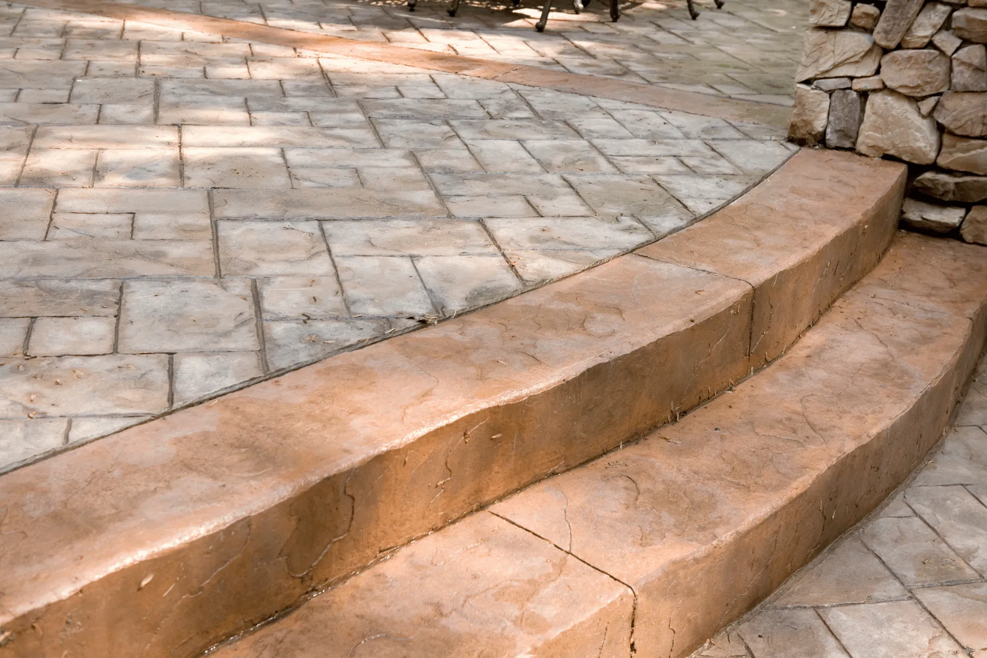 A curved set of outdoor steps with a stone veneer wall at the top right. The steps and the surrounding patio area are made of stamped concrete with a stone-like pattern and texture. Sunlight filters through the scene, casting shadows on the ground.