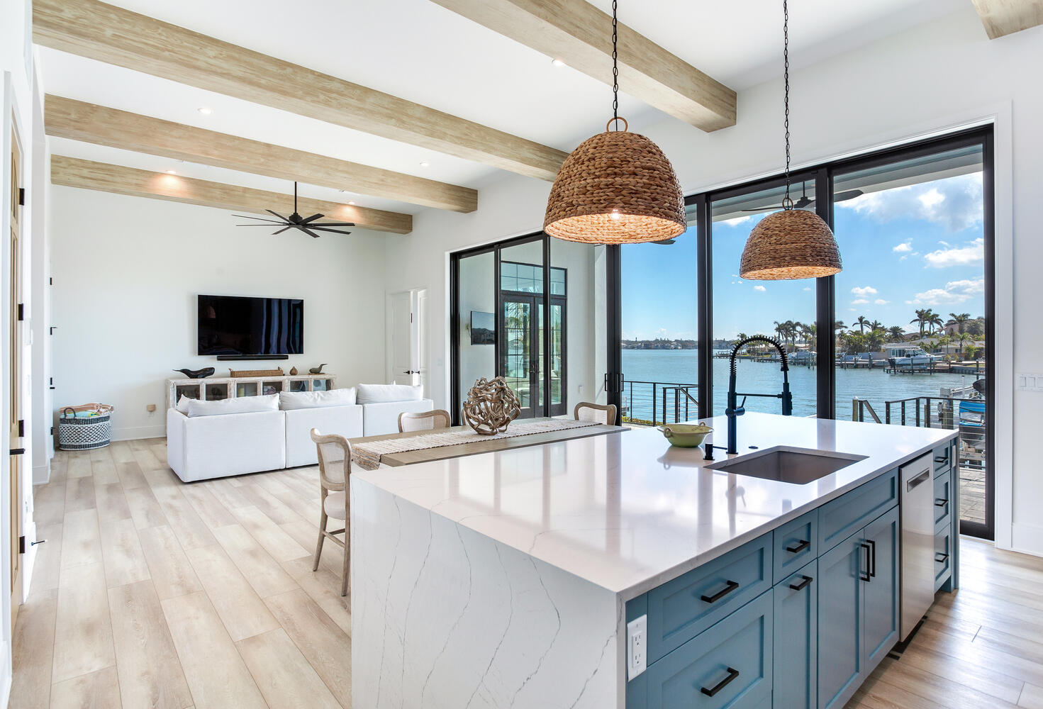 Open-concept AVENTIS kitchen and living area featuring a large island, vaulted ceiling beams, woven pendant lights, natural light, and modern coastal finishes.