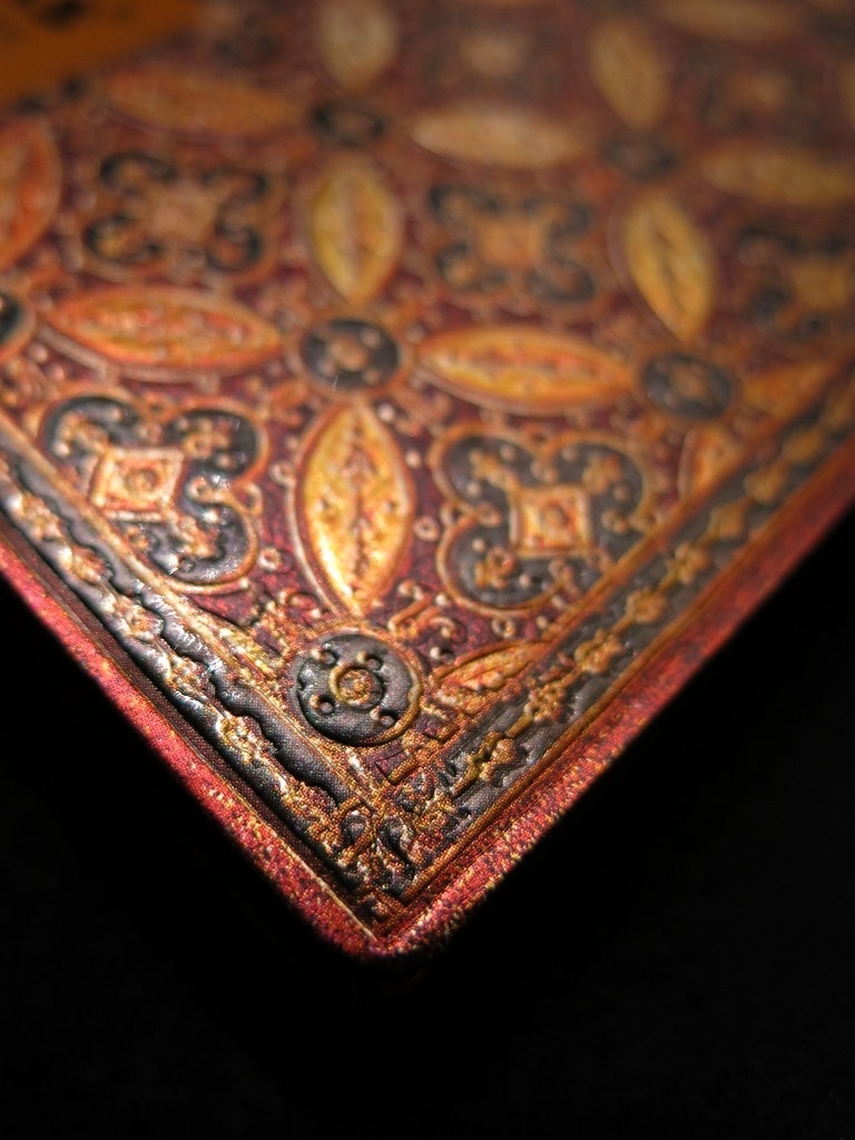 An exceptional close-up product image by Tabula Rasa Social Stationers, showcasing the intricate craftsmanship of a Paperblanks journal cover. The photograph highlights a segment of the cover, adorned with a deeply embossed, ornate pattern in rich gold, red-orange, and black tones. This detailed view perfectly illustrates Paperblanks' dedication to combining artistry with craftsmanship, featuring designs inspired by diverse cultures and art from around the world. The image emphasizes the premium quality and attention to detail synonymous with Paperblanks journals.