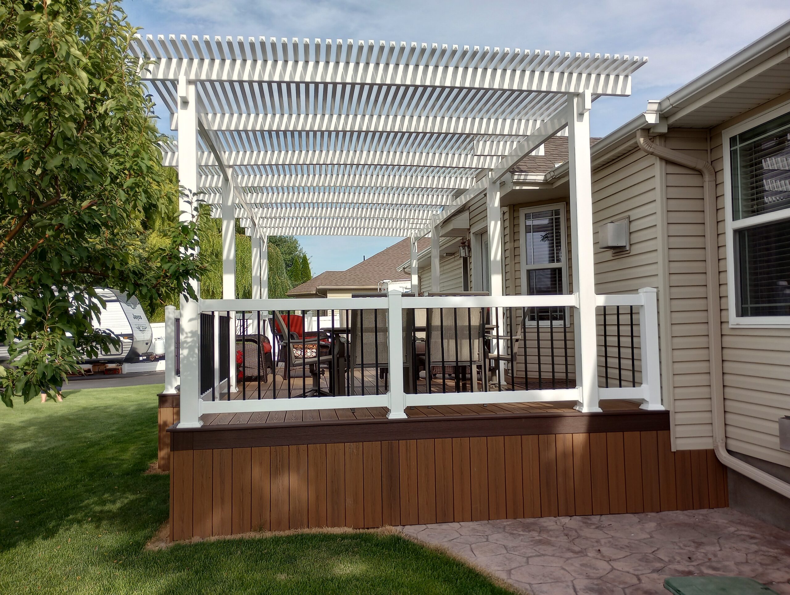 Transform your backyard into a perfect retreat with this custom composite deck and pergola from DeckPro LLC. Enjoy low-maintenance living with durable brown composite decking and a stylish white vinyl pergola that provides refreshing shade. The elegant white railings with black balusters and wide stairs leading to a stamped concrete patio complete this beautiful outdoor living space.