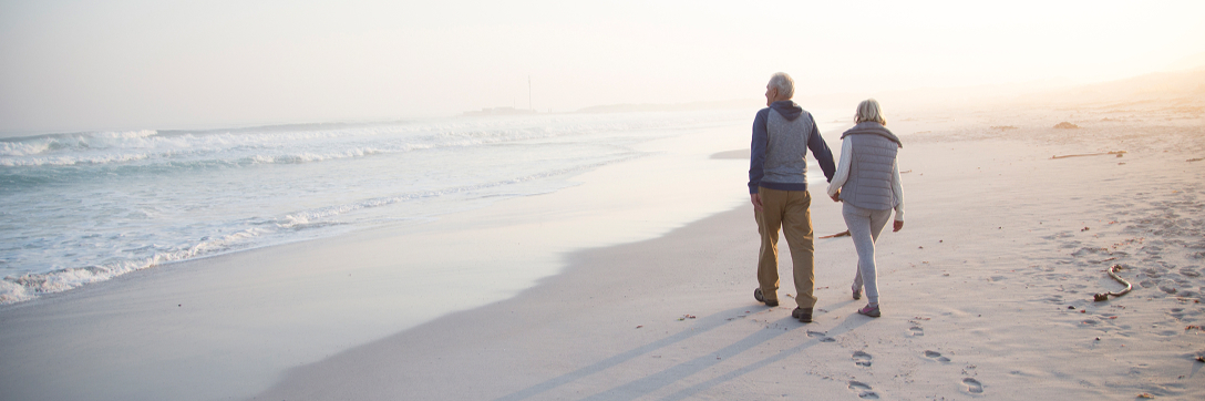An elderly couple walks hand in hand along a serene beach at sunrise, casting elongated shadows on the sand. Waves gently lap the shore, creating a peaceful atmosphere.