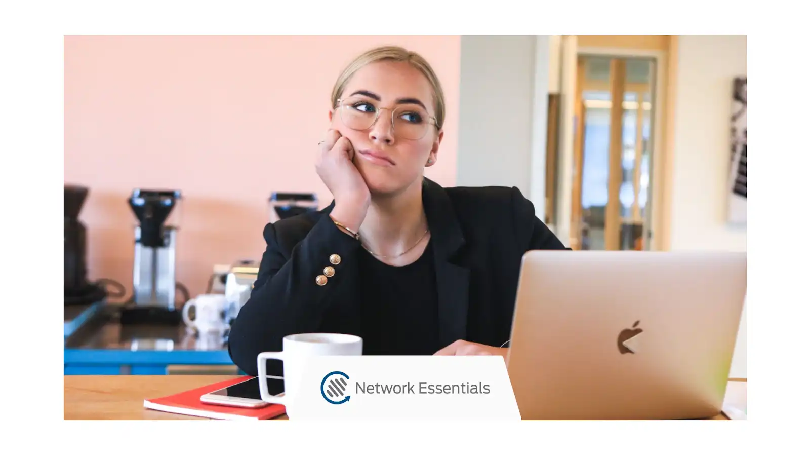 A young woman with blonde hair and glasses sits at a desk with a laptop, looking bored. She is wearing a black blazer over a black shirt and has her hand on her chin. In front of her are a white mug, a red notebook, and a smartphone. A logo for "Network Essentials" is visible. Behind her is a coffee machine. The background appears to be an office setting with light pink walls and glass doors.