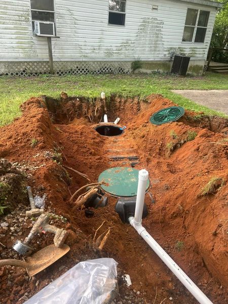 This photograph captures the installation or repair of a residential septic system. It shows a deep excavation in red clay soil revealing two green tank lids and connected white PVC piping.
