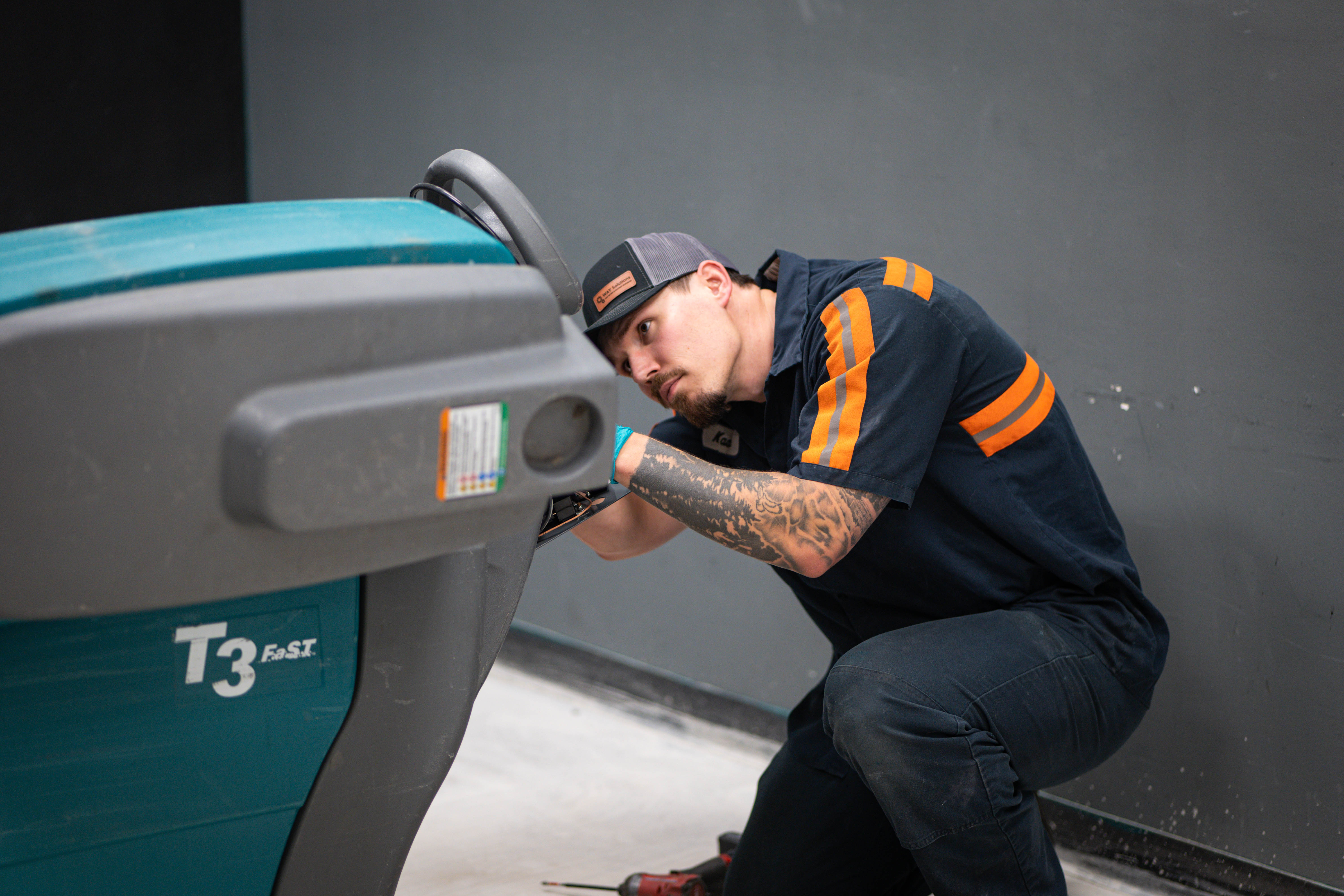 A certified WaySolutions technician closely inspects the side panel and mechanical linkages of a trusted teal Tennant T3 walk-behind floor scrubber. Providing expert commercial cleaning equipment maintenance in Layton, Utah, our team ensures your facility's daily workhorses remain reliable shift after shift.