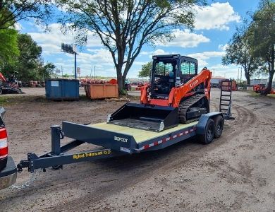 A skid steer loader is secured on a flatbed trailer attached to a pickup truck in an outdoor work area. The setup highlights equipment transport for construction or landscaping jobs, showing proper loading and hauling practices.
