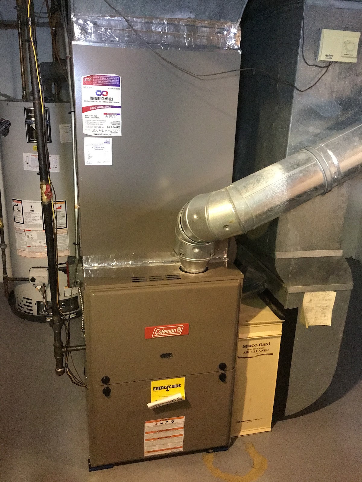 A residential furnace and water heater are installed in a utility room. The furnace is a brown Coleman brand unit with an "Energyguide" sticker. A large silver air duct connects to the top of the furnace. To the left is a silver water heater with various pipes and wiring attached. A cream-colored "Space-Gard" air cleaner unit is next to the furnace. The room has a concrete floor with a yellow horseshoe-shaped marking.