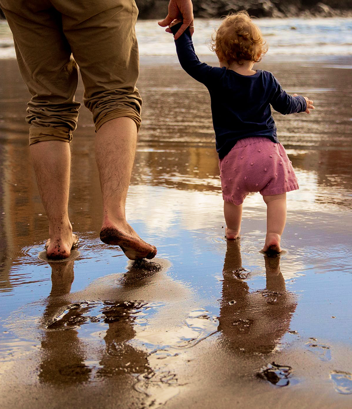 A father walking hand-in-hand with his daughter on the beach.