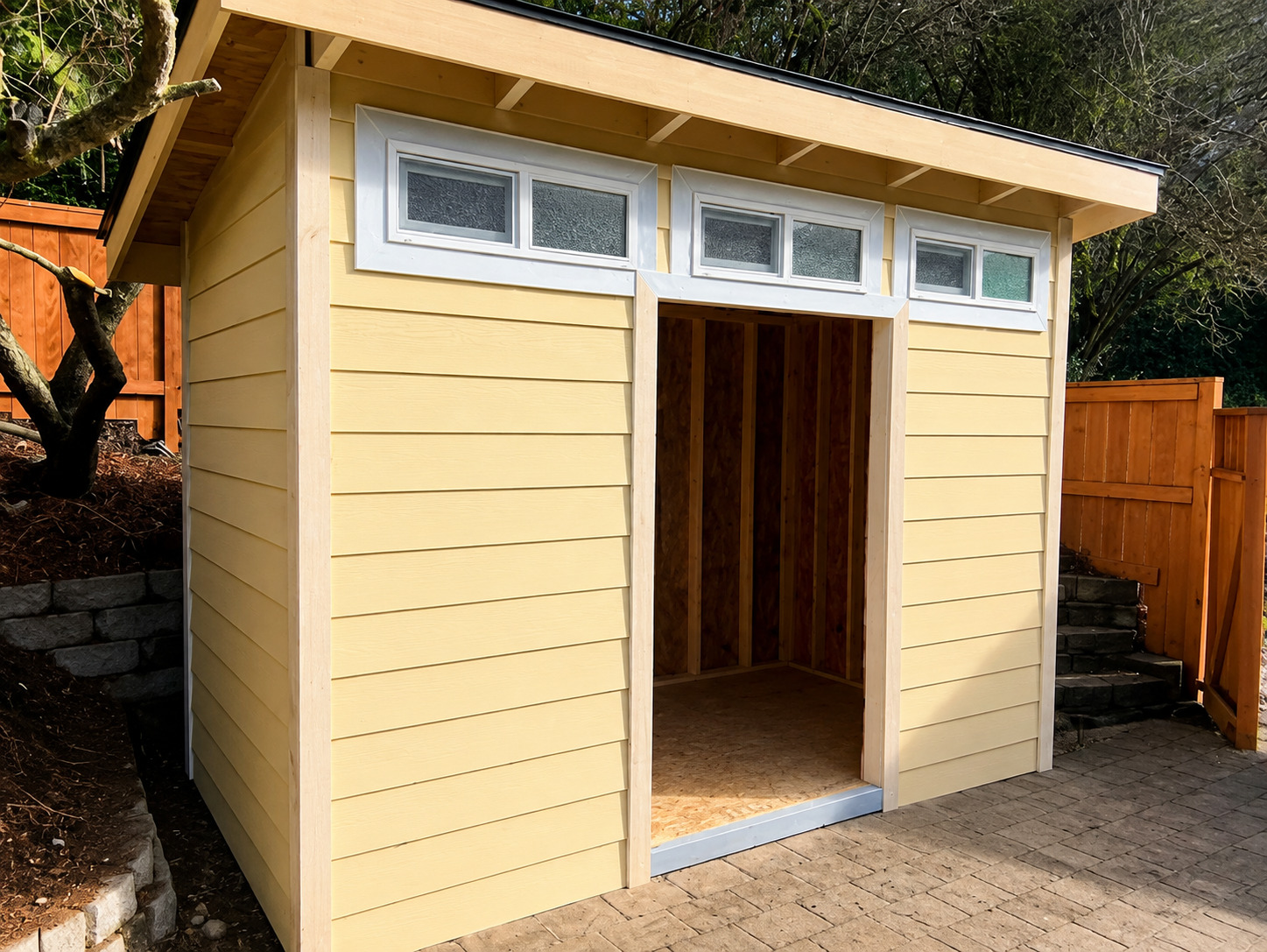 A compact backyard shed features pale yellow horizontal siding, white-trimmed windows, a wide open d 