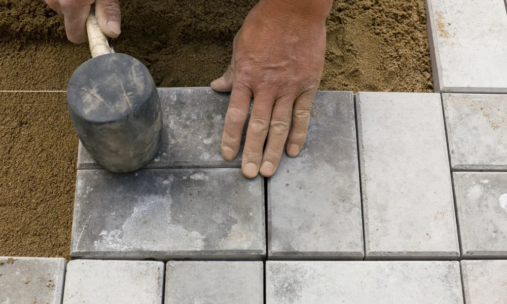 A close-up shot shows a person's hands laying grey rectangular paving stones on a bed of sand. One hand is holding a rubber mallet to tap a stone into place, while the other hand is positioned to guide the stone. The paving stones are arranged in a pattern, and a thin string is visible in the sand, likely used as a guideline.