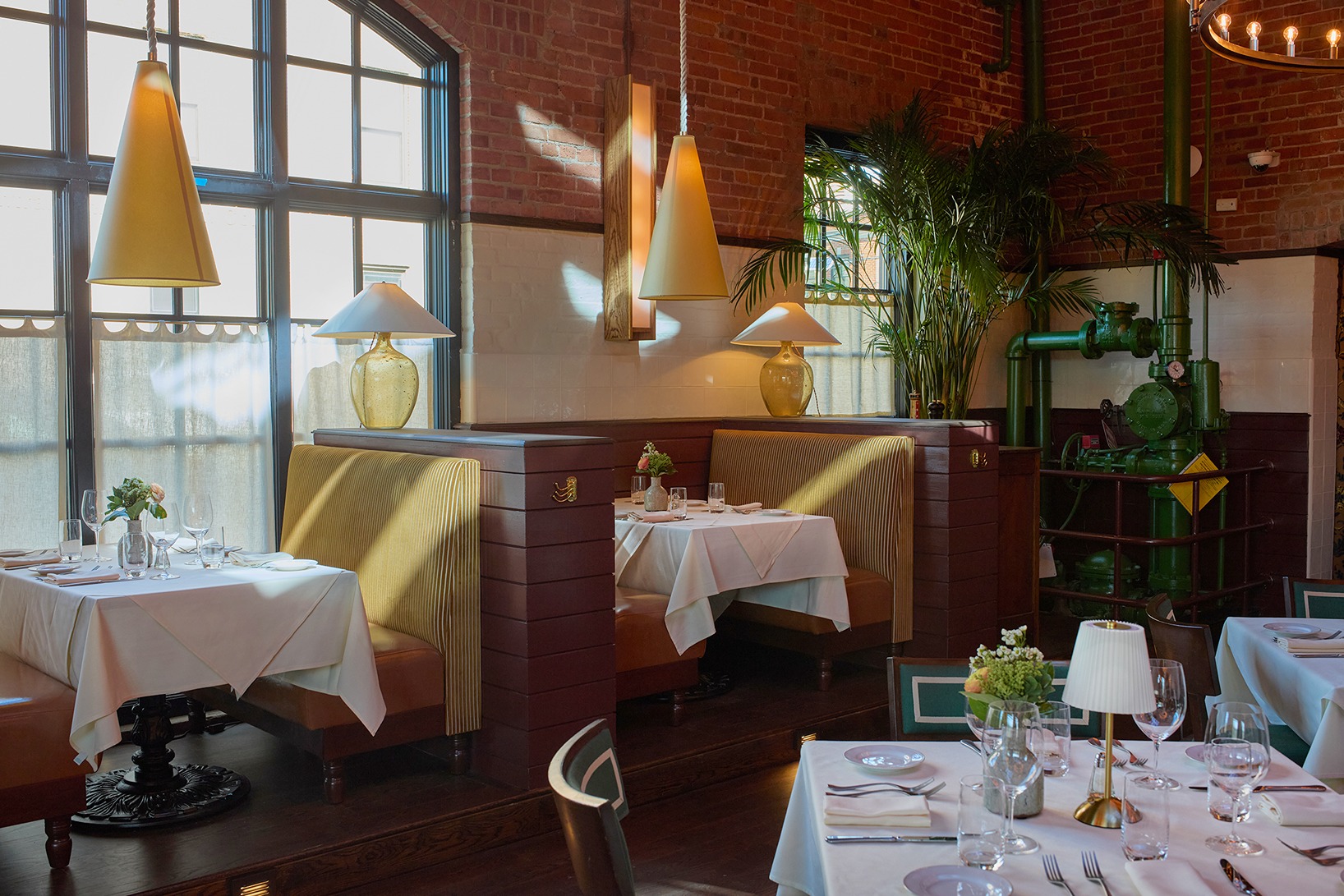 Sun-drenched dining booths with striped cushions are set against white and red brick walls. Large industrial windows with cafe curtains let in bright afternoon light, highlighting the elegant table settings.