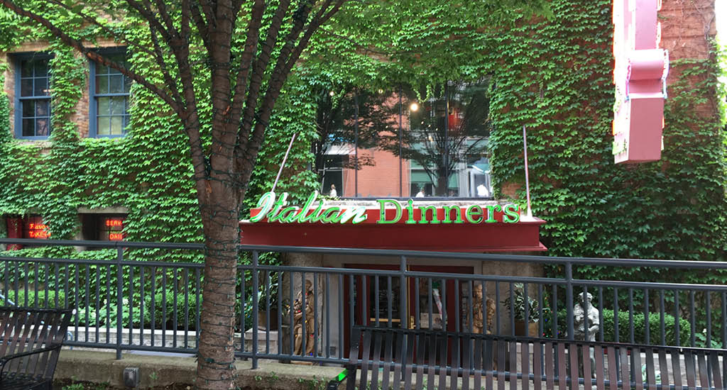 The exterior of Buca di Beppo Columbus showing greenery surrounding the building and windows with an Italian Dinners sign.