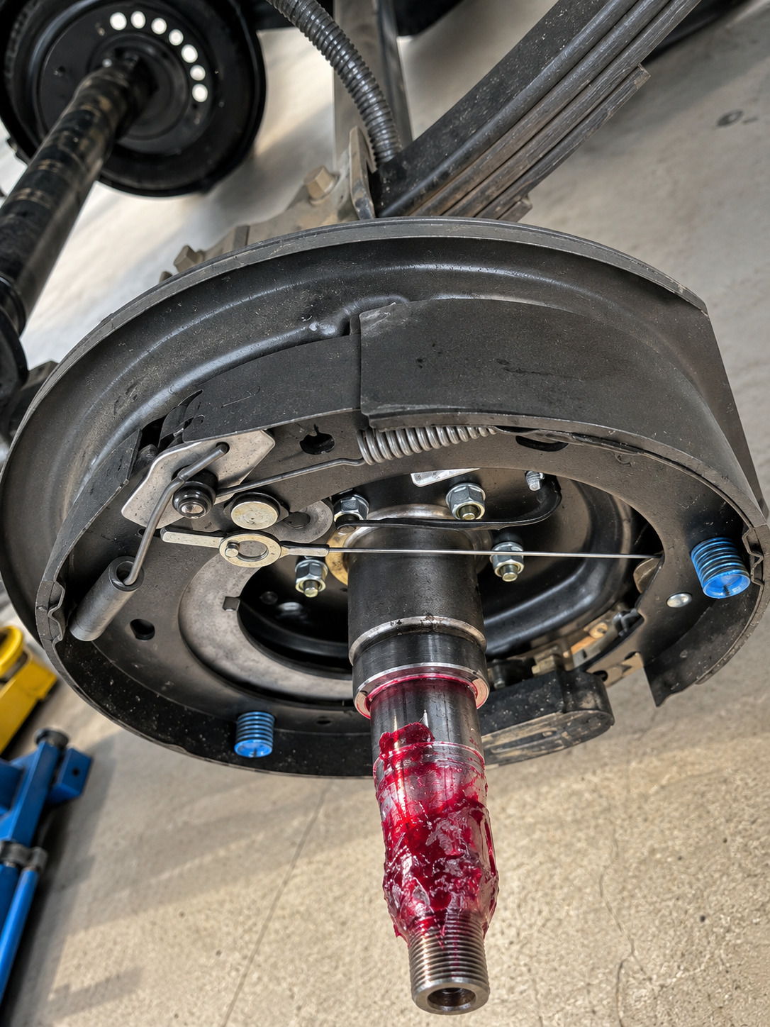 Close-up of an RV drum brake assembly with exposed brake shoes, springs, adjuster hardware, axle spindle, red grease, mounting plate, and suspension components visible during maintenance inside a repair workshop.