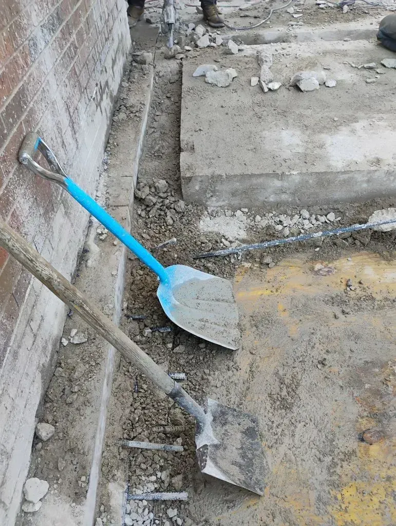 Shovels placed near partially removed concrete slab beside brick wall, with exposed soil, debris, an 