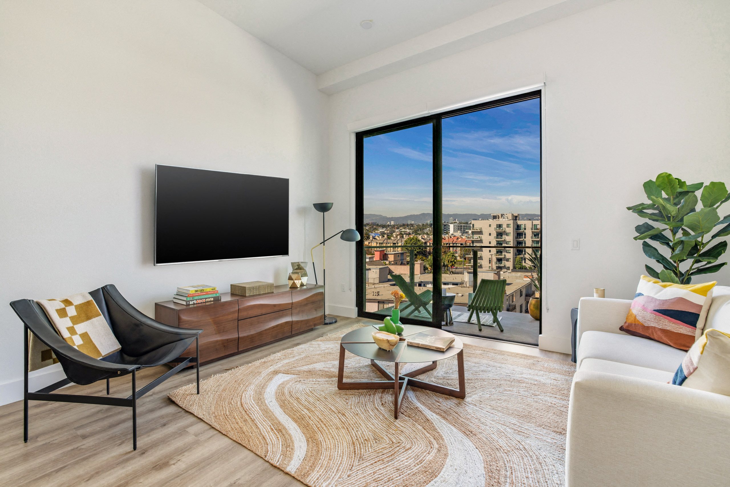 A living room with a black chair, a white sofa, a wooden coffee table, and a large window with a view of the city.