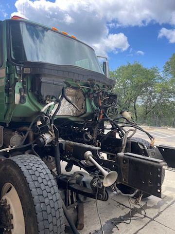Front section of a heavy-duty green truck with hood removed, exposing engine bay, wiring, hoses, frame components, steering hardware, and front wheel while parked outdoors beside trees and pavement nearby.