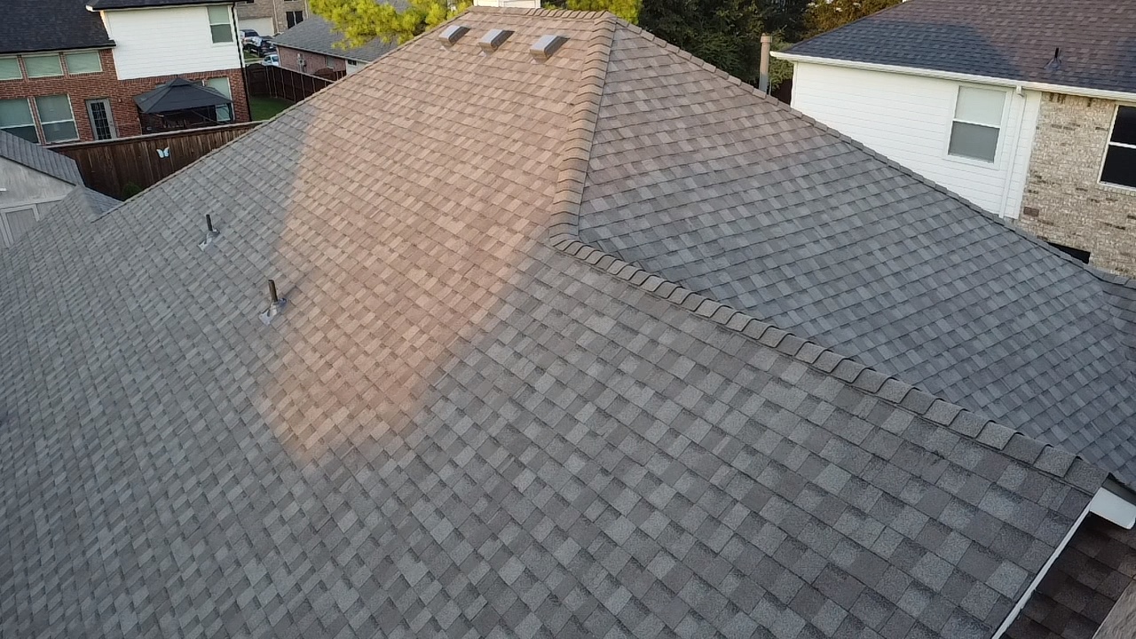 An aerial view of a residential rooftop covered in gray asphalt shingles. The roof has a complex structure with multiple peaks and valleys, and several vents protrude from the surface. The sunlight casts shadows across the shingles, highlighting their texture. In the background, parts of neighboring houses and trees are visible, suggesting a suburban setting. The overall impression is of a well-maintained roof in an outdoor environment.