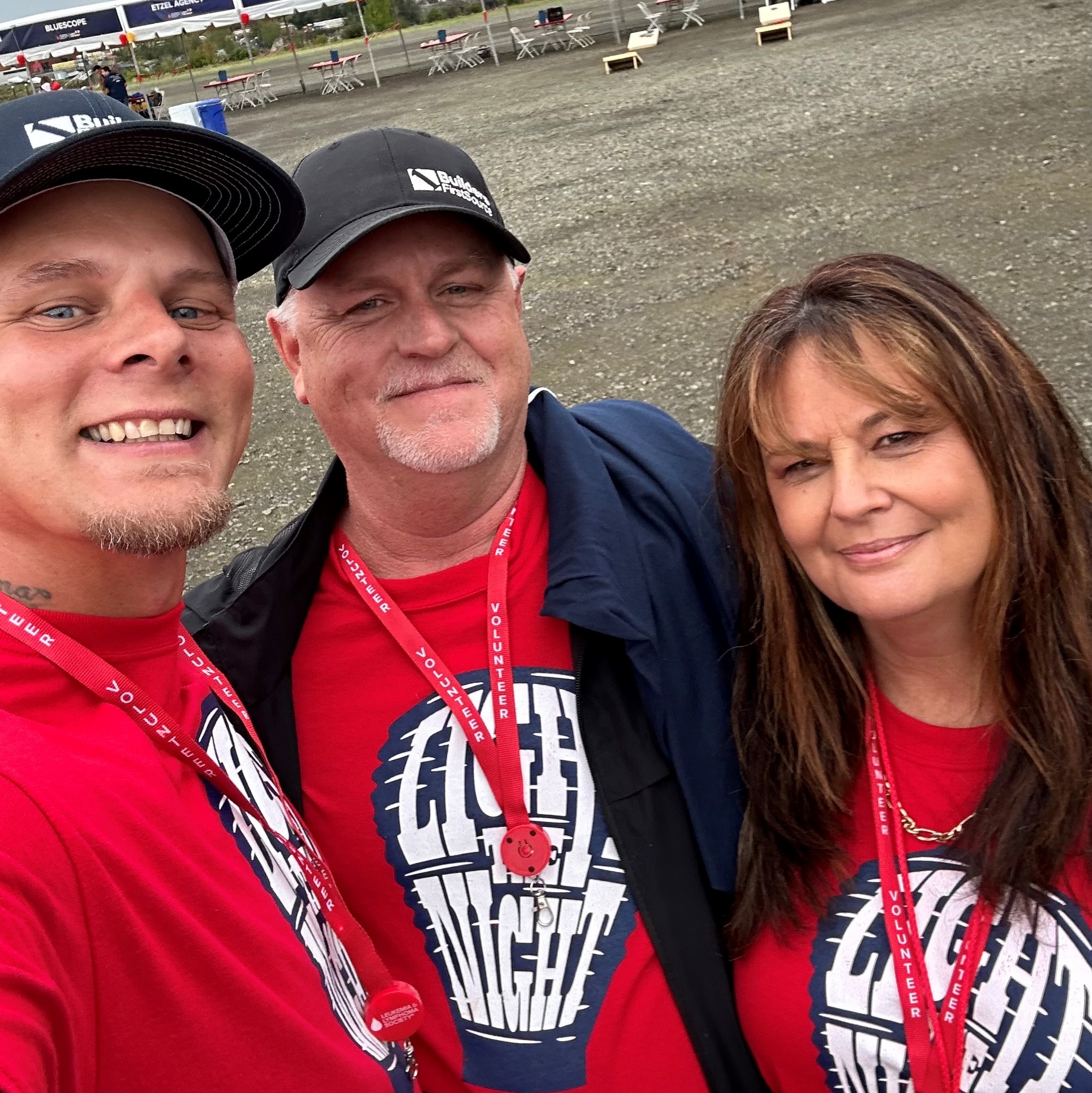 A selfie of three people smiling outdoors, all wearing matching red "Light the Night" t-shirts. They have red lanyards around their necks that say "Volunteer." The man on the left is wearing a black baseball cap, and the man in the middle is wearing a "Builders FirstSource" cap and a jacket. They are standing on a gravel area with some tables and fencing visible in the background, likely at an outdoor event or charity walk.