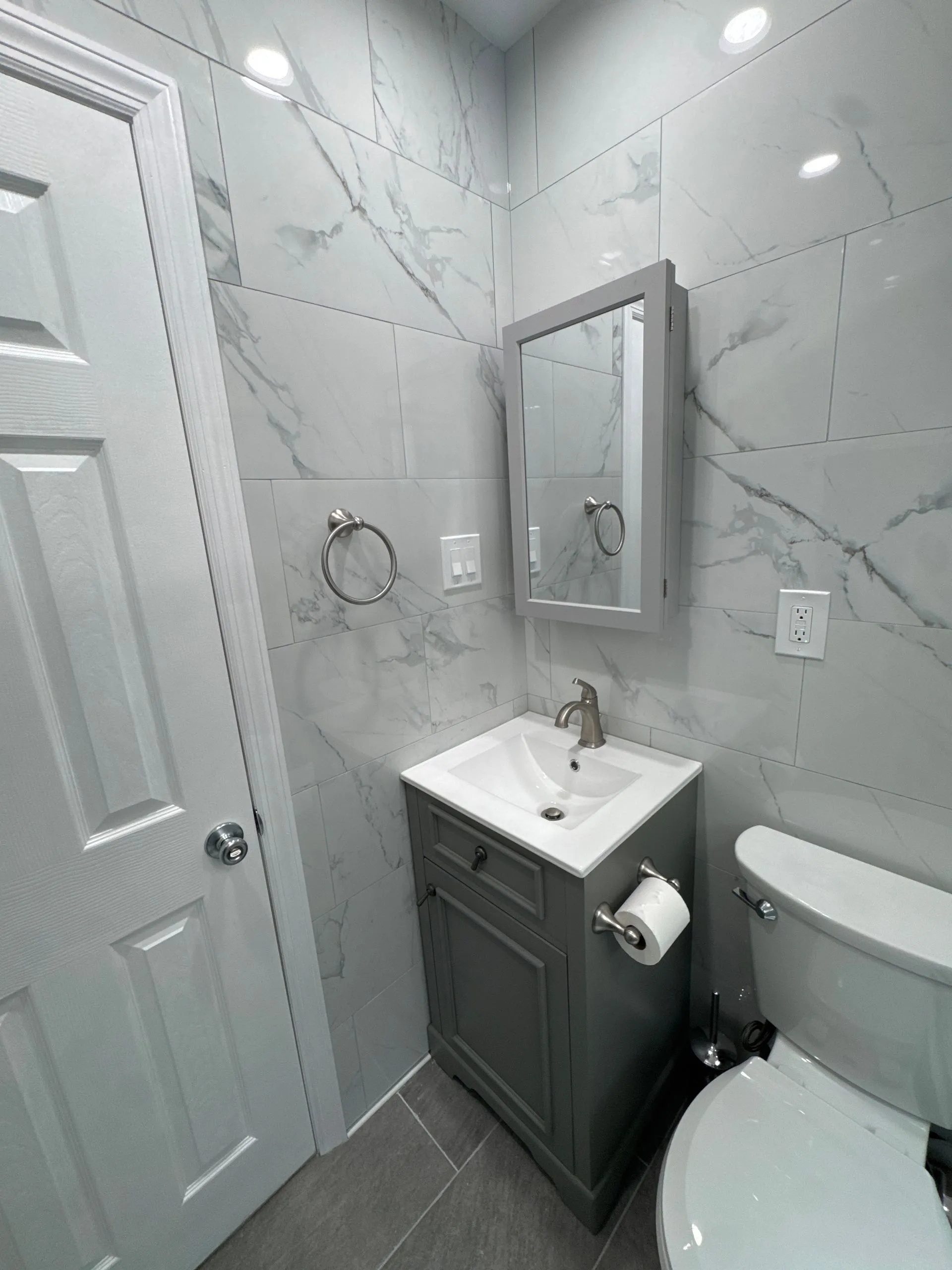 Modern bathroom renovation featuring marble-look wall tiles, gray vanity cabinet, recessed lighting, and contemporary fixtures. The design maximizes style, functionality, and efficient use of space.