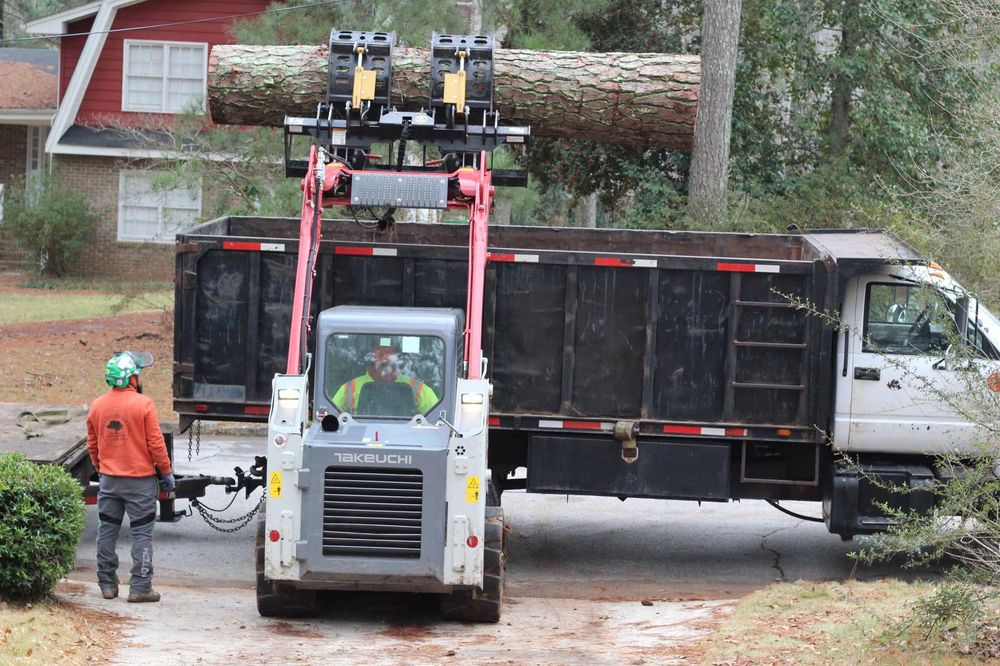 Efficient debris removal at a residential site. A compact loader carefully maneuvers a heavy log into a disposal truck, supported by a professional ground crew ensuring a clean workspace.