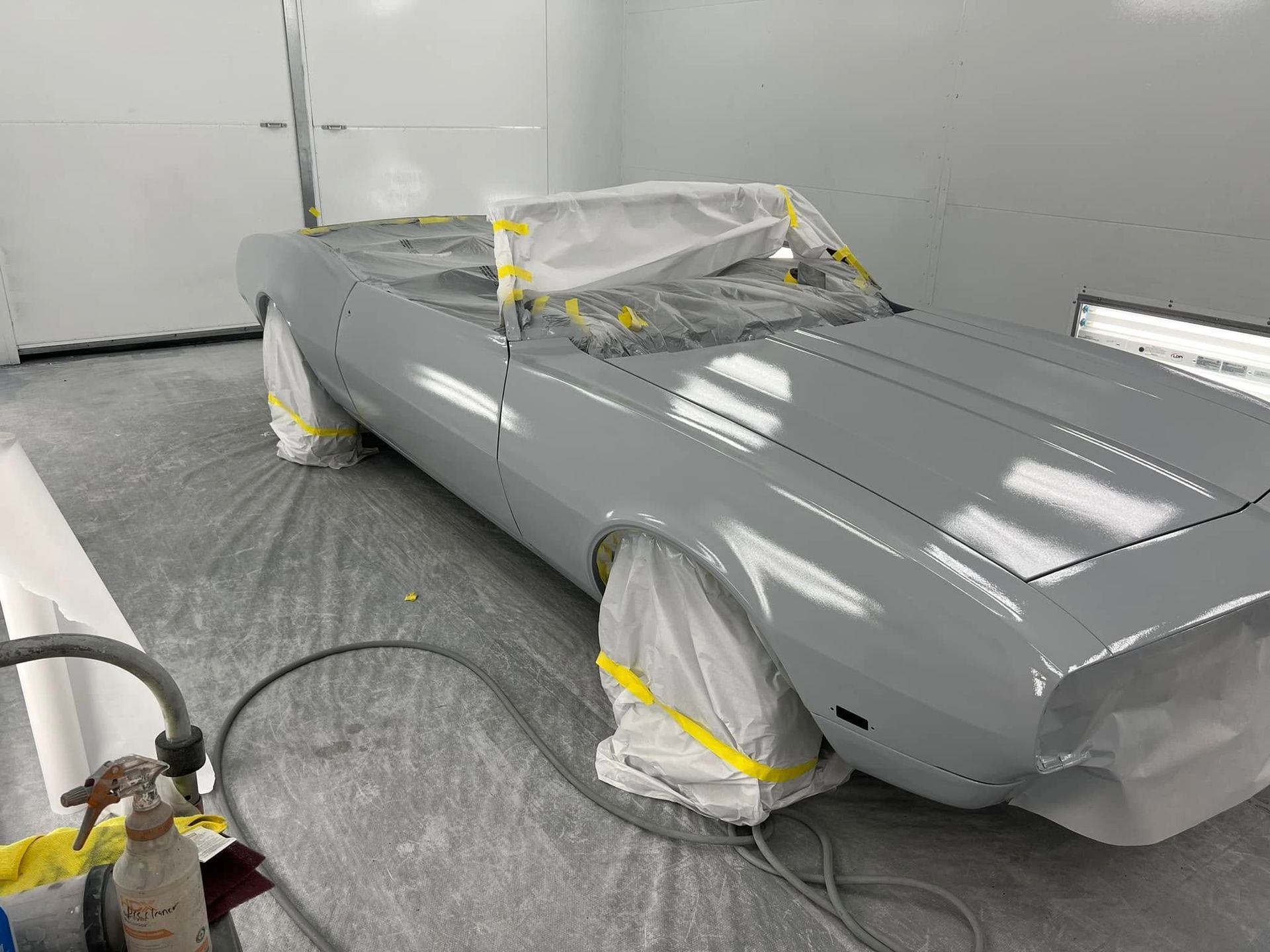 A restored classic convertible receives primer coating before color application. Protective masking covers key components while technicians prepare the vehicle for its final paint finish.