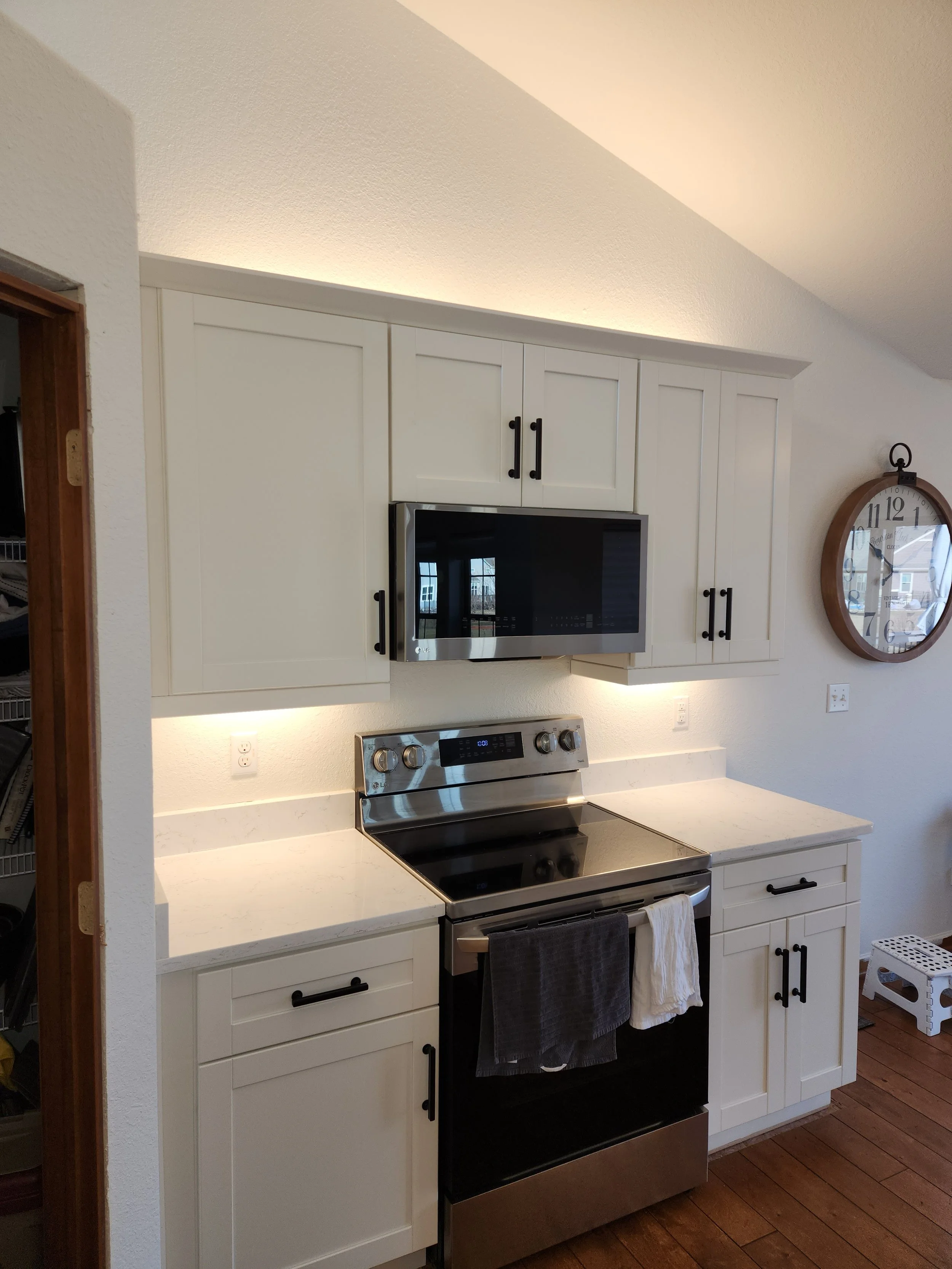 A modern kitchen with white cabinets, a stainless steel range, and a microwave above it. A large round clock with a wooden frame hangs on the wall to the right. The countertops are white and speckled. The flooring is dark wood. Under-cabinet lighting adds a warm glow.