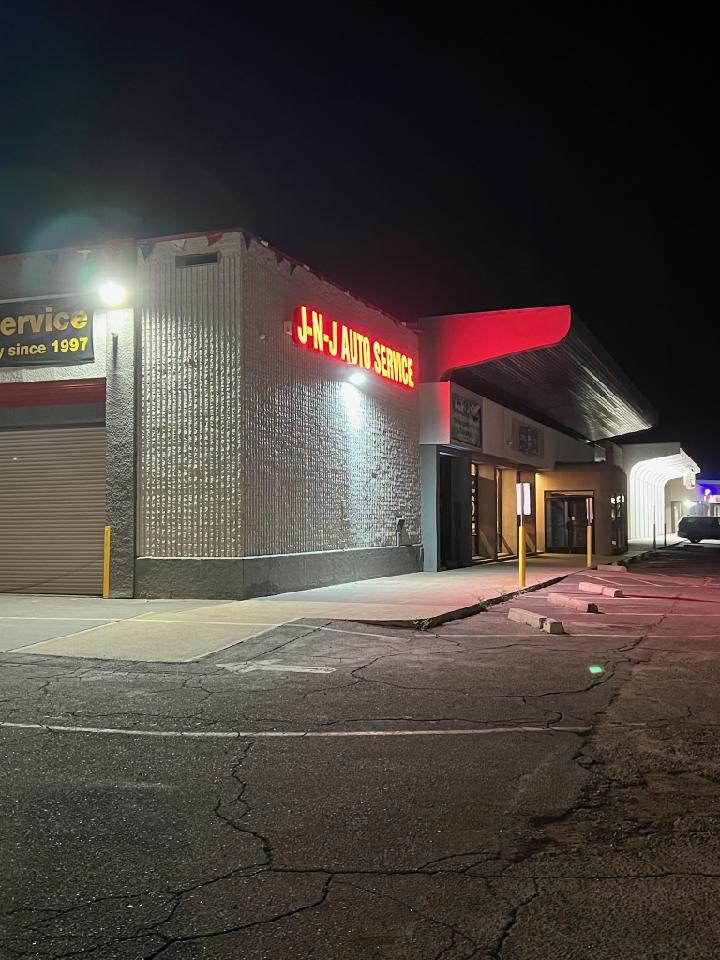 A nighttime exterior view of J-N-J Auto Service, with red neon signage prominently displayed on the building's facade. The building has a corrugated metal exterior and a large garage door on the left. The parking lot in front is paved and cracked, with concrete parking barriers visible.