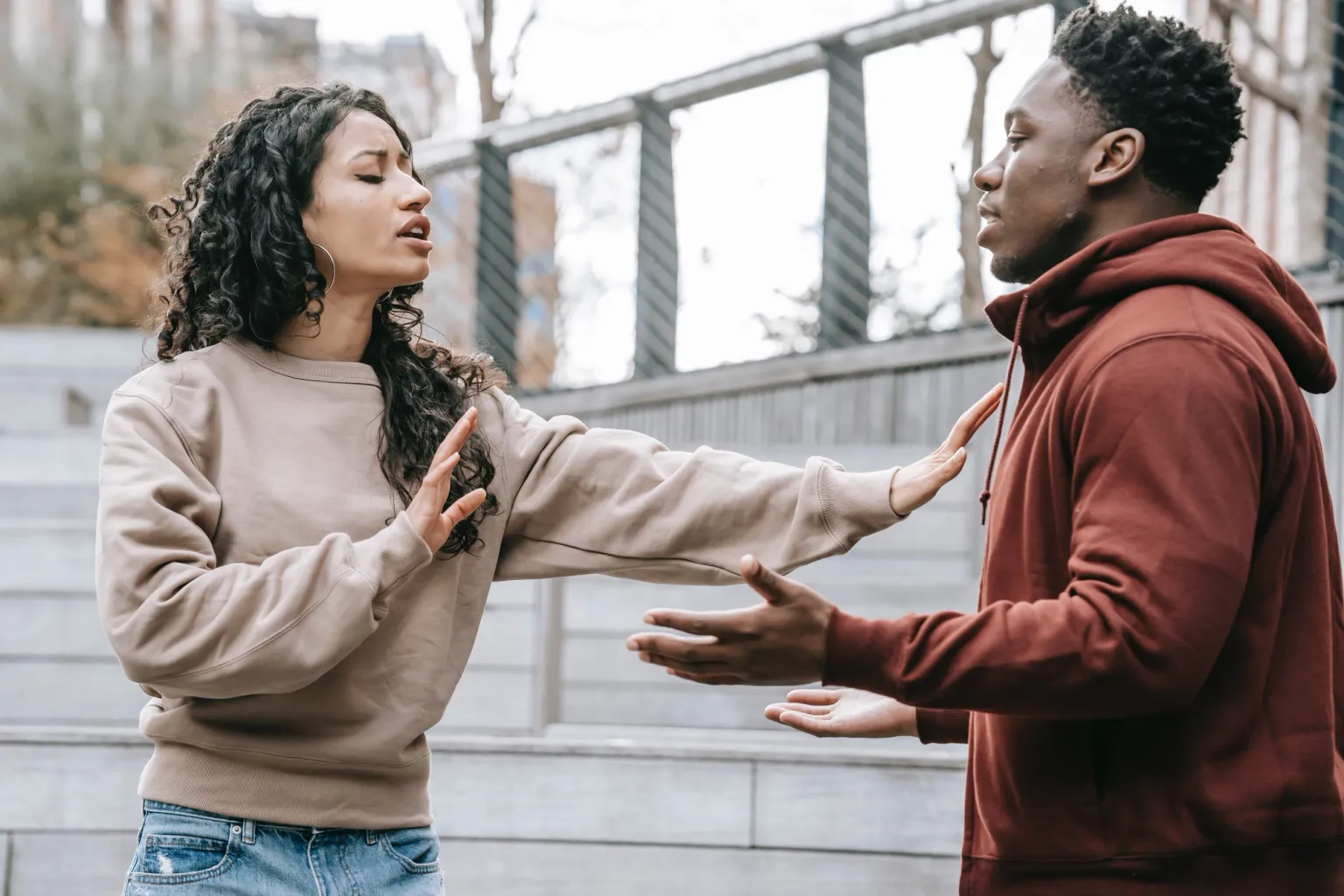 A man and a woman appear to be in the middle of an argument, with the woman holding up her hands in a defensive gesture and looking away from the man. The man's hands are open and outstretched towards her.