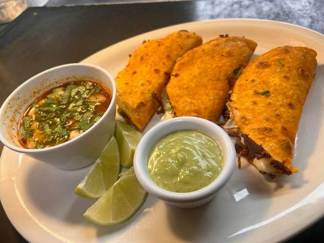 Three crispy birria tacos filled with slow-cooked beef and melted cheese, served with consommé, lime wedges, and salsa at Catramex Restaurant.