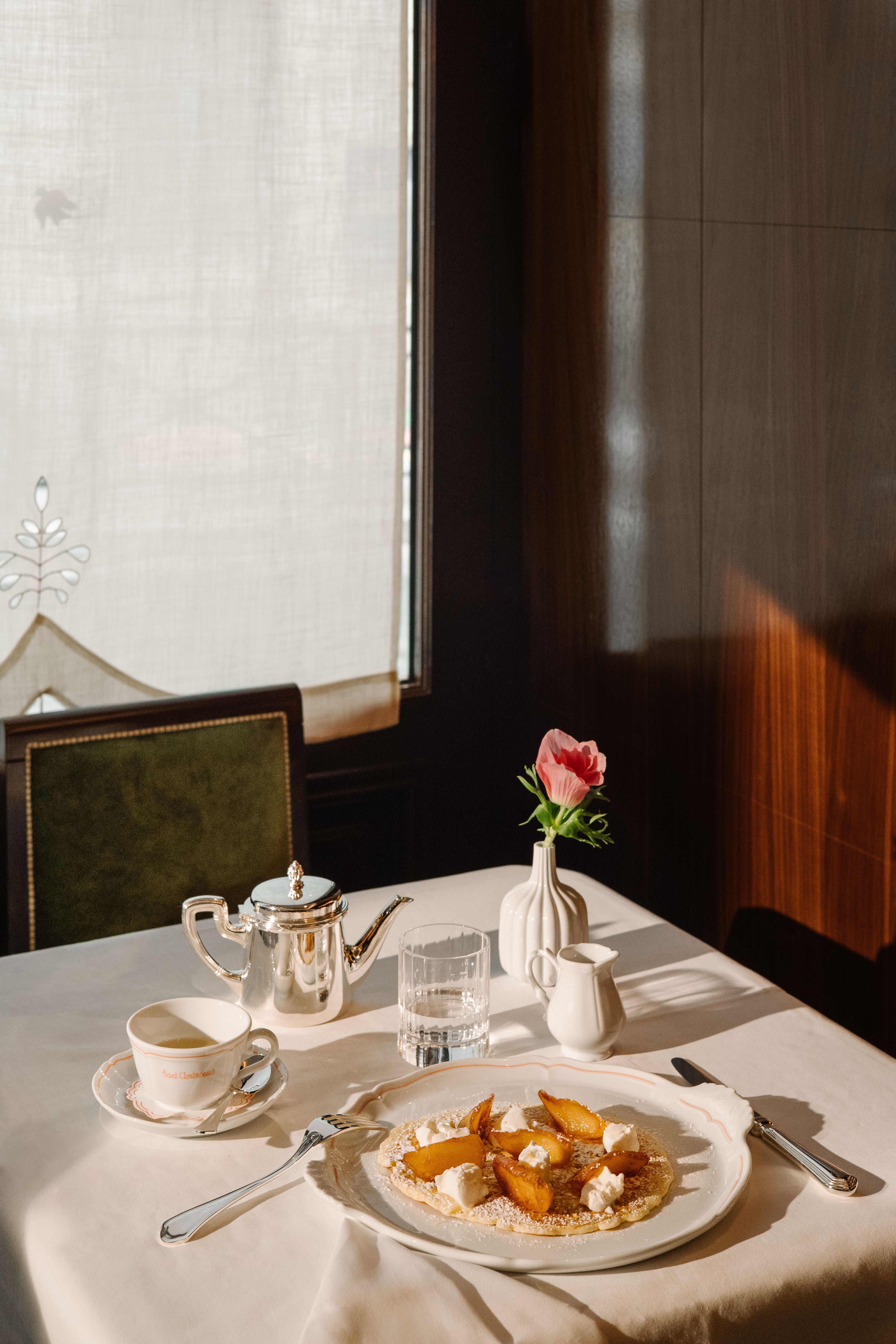 Sunlit café table set with a white tablecloth, featuring a plated pancake topped with caramelized fruit and dollops of cream, alongside a silver teapot, coffee cup and saucer, water glass, small milk pitcher, and a single pink flower in a vase by a window.