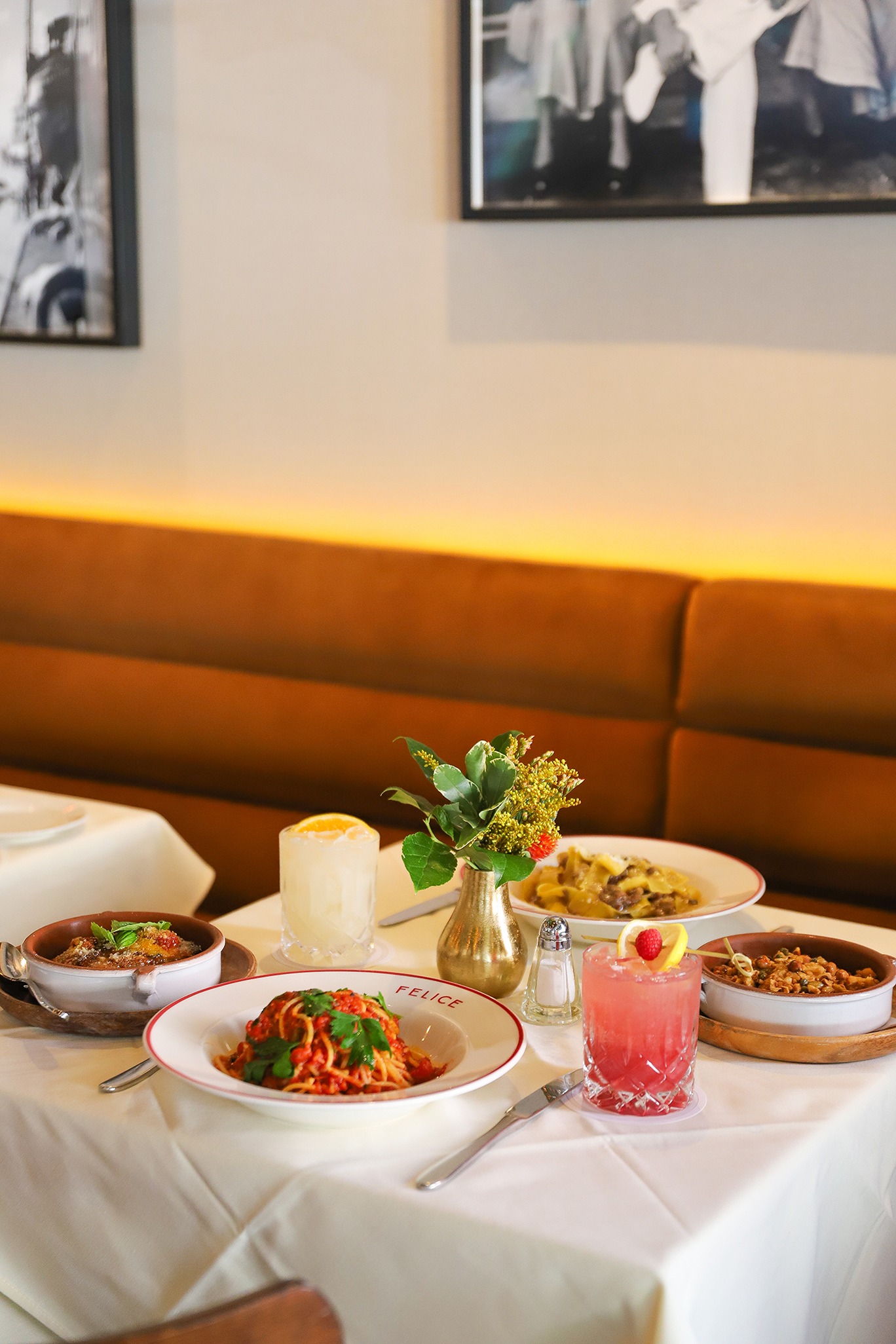 A white-clothed table featuring spaghetti pomodoro, rigatoni, and vibrant cocktails. The background shows a comfortable brown leather banquette and framed black-and-white photography.