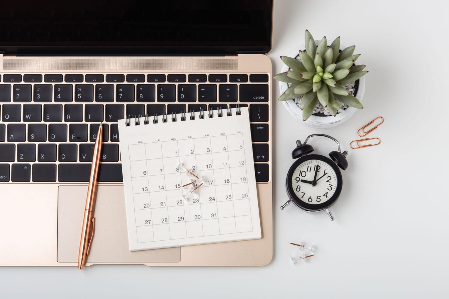 A top-down view of a desk with a laptop, a spiral-bound calendar, a rose gold pen, a succulent plant, and a vintage alarm clock. The calendar is open to the month of March, with push pins marking the 9th, 15th, and 17th. The desk surface is white, and the overall aesthetic is clean and organized, suggesting a workspace.