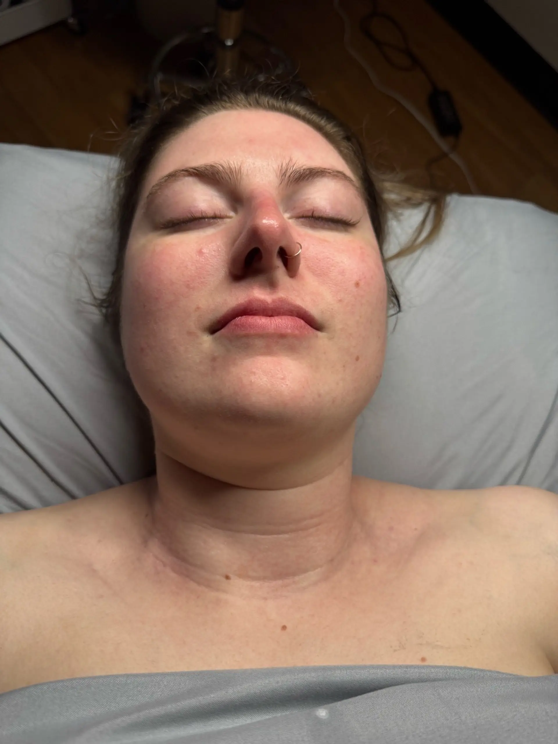 A close-up overhead shot of a person lying down with their eyes closed and a septum piercing.