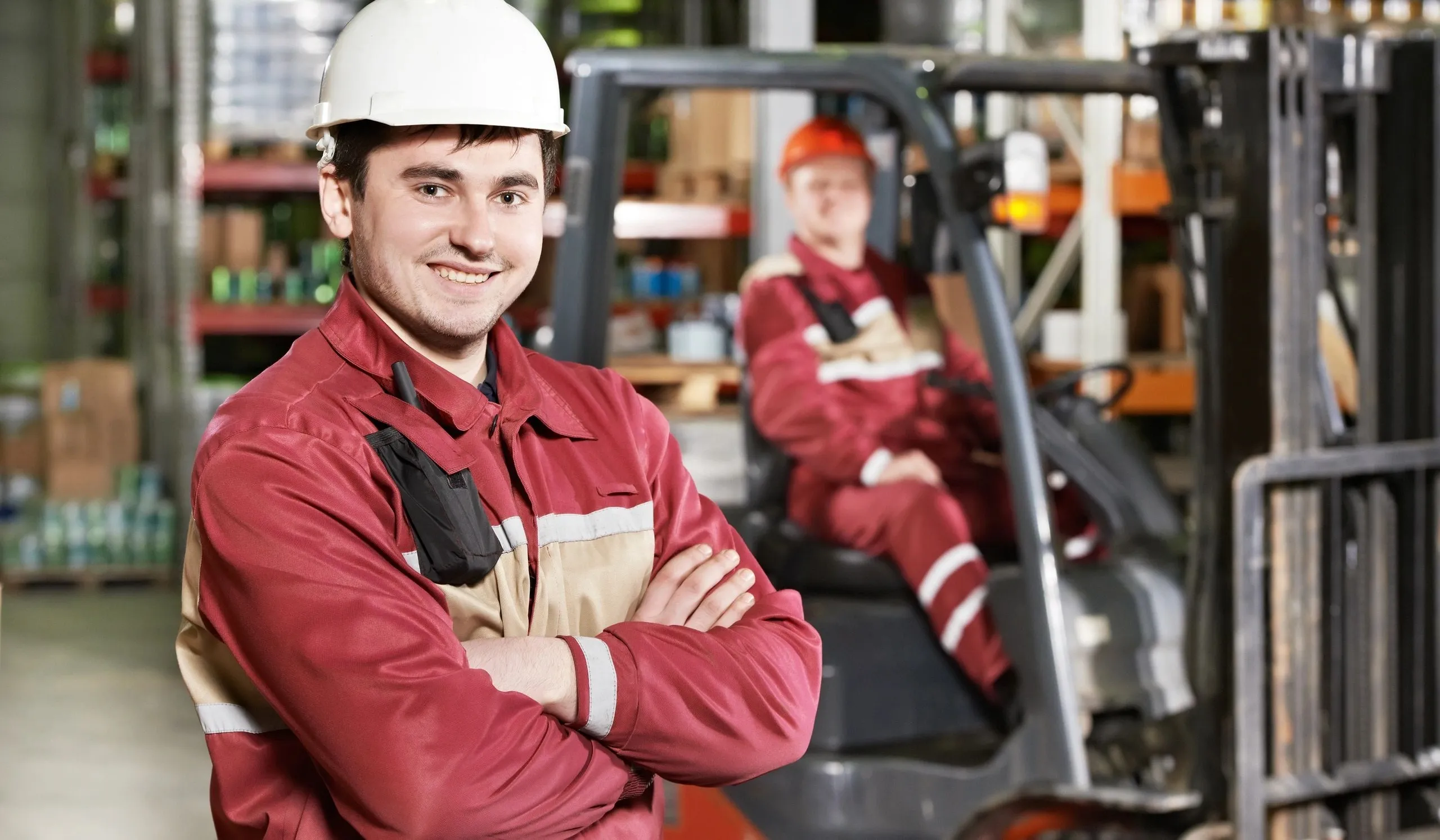A man in a red uniform and white hard hat smiles with his arms crossed in front of a forklift and another worker in the background, suggesting a warehouse or industrial setting. The man in the foreground is looking directly at the camera, appearing friendly and approachable. The background shows shelving units filled with products, typical of a storage or distribution facility.